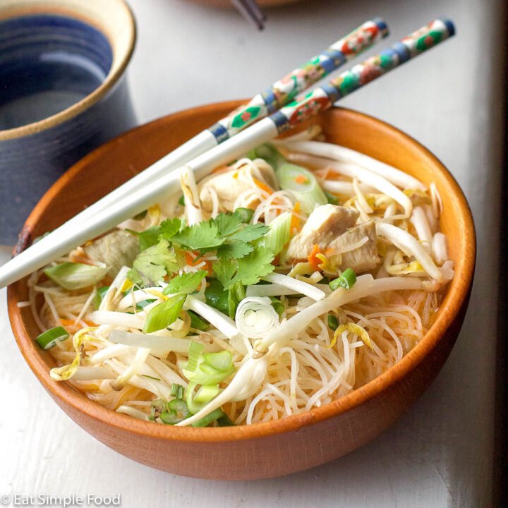 2 wood bowls and one pair of chopsticks. Bowls have rice noodles and a tan sauce with cilantro, mung beans, and sliced green onion garnish. side top view.
