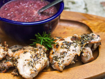 Pan Fried Chicken Thigh with a blue bowl of Grape sauce on a Wood Serving Plate with a sprig of rosemary
