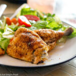 Cooked Chicken Thigh and Leg with A Brown Dry Rub On A white plate with a green salad and red tomato salad.