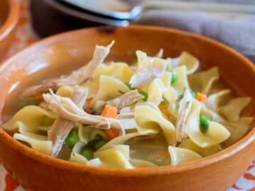 Side view of wood bowl of chicken soup with egg noodles, carrots, and peas.