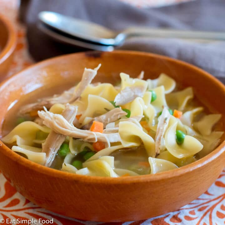 Side view of wood bowl of chicken soup with egg noodles, carrots, and peas.