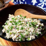 Brown bowl full of cooked diced chicken, diced celery and pickles, diced red onions stirred with white mayonnaise and garnished with parsley. Wood spoon in it.
