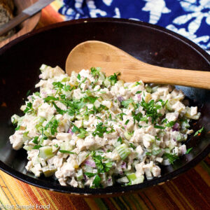Brown bowl full of cooked diced chicken, diced celery and pickles, diced red onions stirred with white mayonnaise and garnished with parsley. Wood spoon in it.