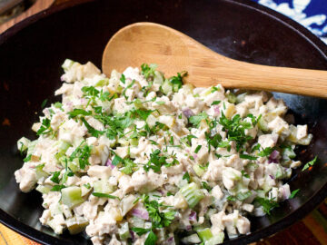 Brown bowl full of cooked diced chicken, diced celery and pickles, diced red onions stirred with white mayonnaise and garnished with parsley. Wood spoon in it.