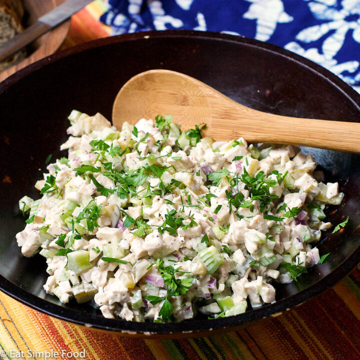 Brown bowl full of cooked diced chicken, diced celery and pickles, diced red onions stirred with white mayonnaise and garnished with parsley. Wood spoon in it.