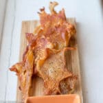 6 crispy roasted chicken thigh and leg skins on a small rectangle wood cutting board with an orange square small serving dish of hot sauce.