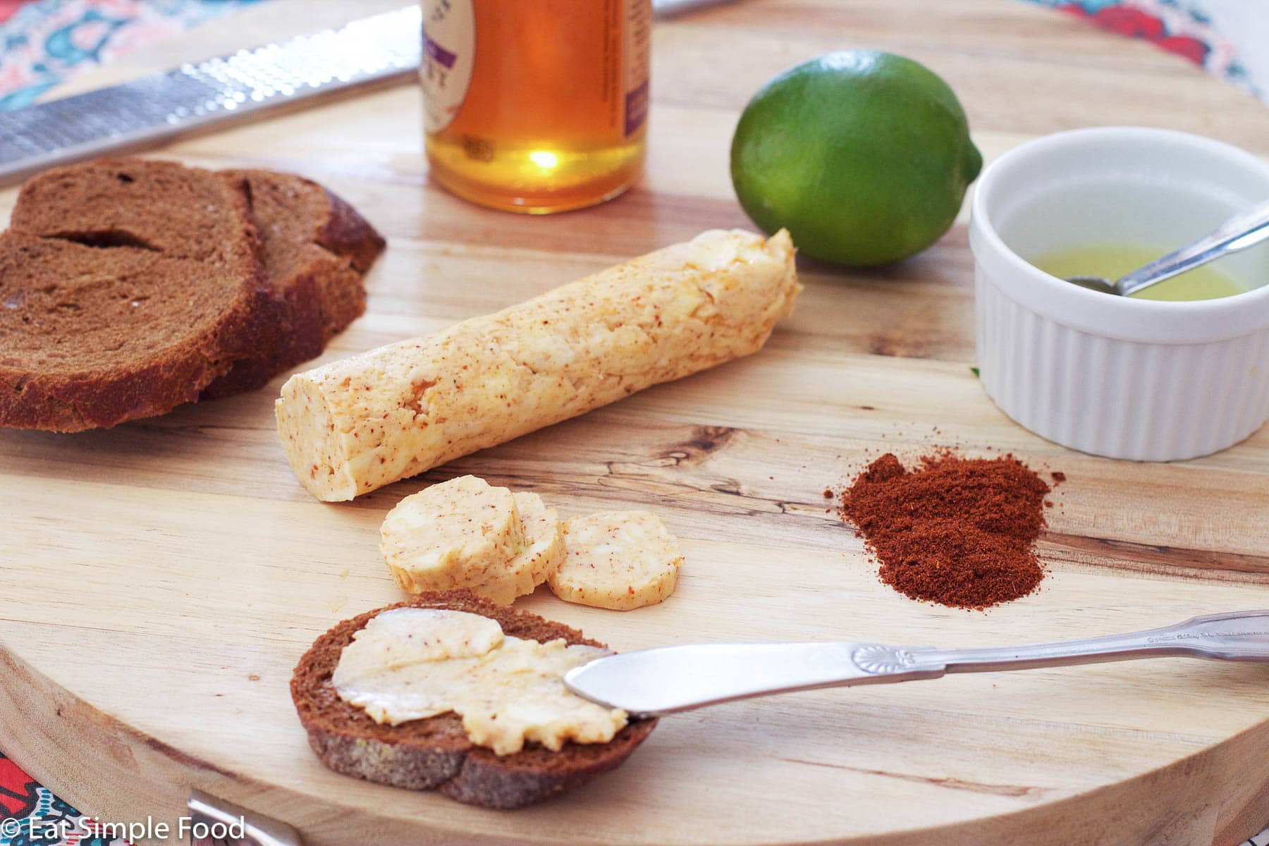 Chili Lime Compound Butter Roll on a wooden cutting board with a bowl of fresh lime juice with a teaspoon in it, fresh sliced bread, a lime, and ground chili powder with a butter knife.