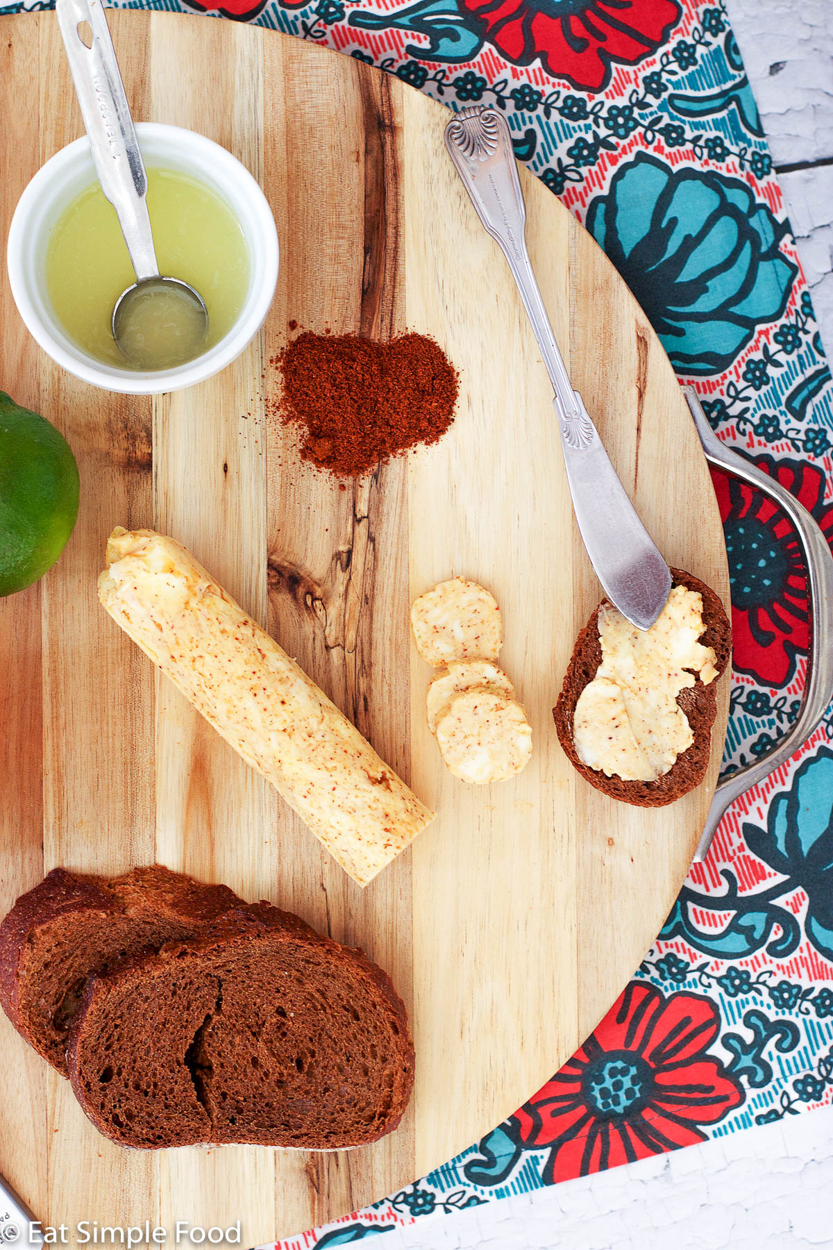 Chili Lime Compound Butter Roll on a wooden cutting board with a bowl of fresh lime juice with a teaspoon in it, fresh sliced bread, a lime, and ground chili powder with a butter knife. horizontal view