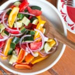 White bowl of chunky red onions, cucumbers, tomatoes, yellow and red peppers salad. Wood spoon it it.
