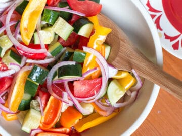 White bowl of chunky red onions, cucumbers, tomatoes, yellow and red peppers salad. Wood spoon it it.