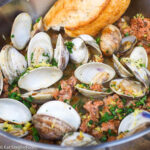 Open cooked clams and chorizo in a steel deep pan. Garnished with lemon zest and parsley. Torn Baguette on side of pan.