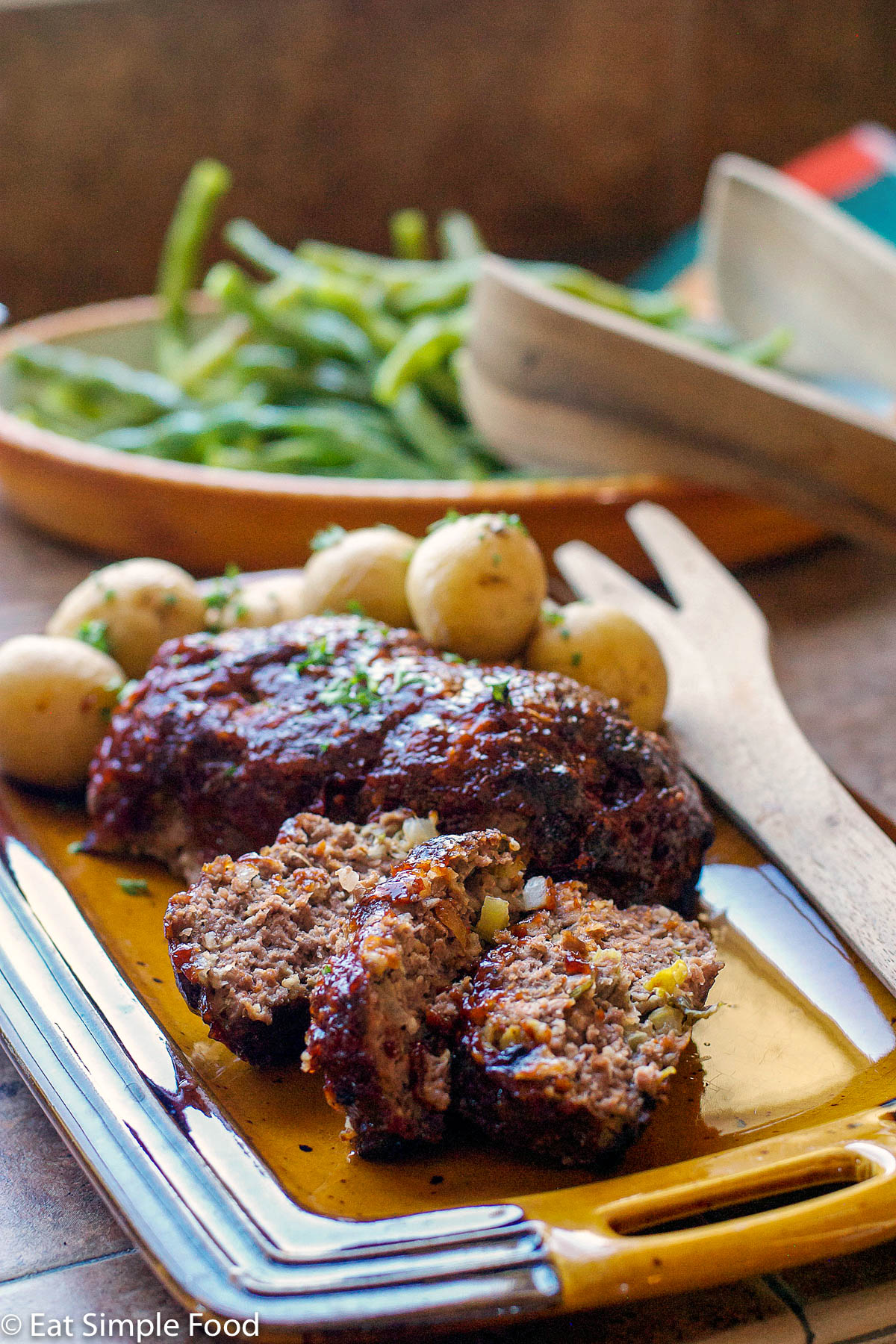Vertical view. Meatloaf on a brown rectangle platter with a couple sliced pieces in front. Baby Baked Potatoes and cooked Green beans in background.
