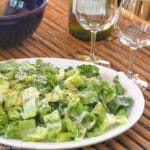 Chunked Romaine Lettuce on a white platter dusted with Parmesan cheese. Tongs on side. 2 glasses of empty wine glasses on side.