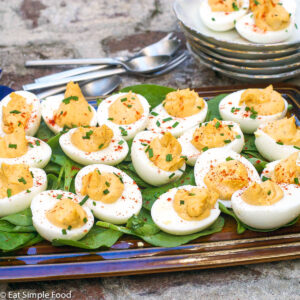 Multiple deviled eggs laying on a bed of spinach on a brown rectangle plate with a chive and paprika garnish.