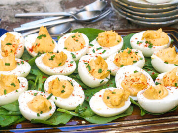 Multiple deviled eggs laying on a bed of spinach on a brown rectangle plate with a chive and paprika garnish.