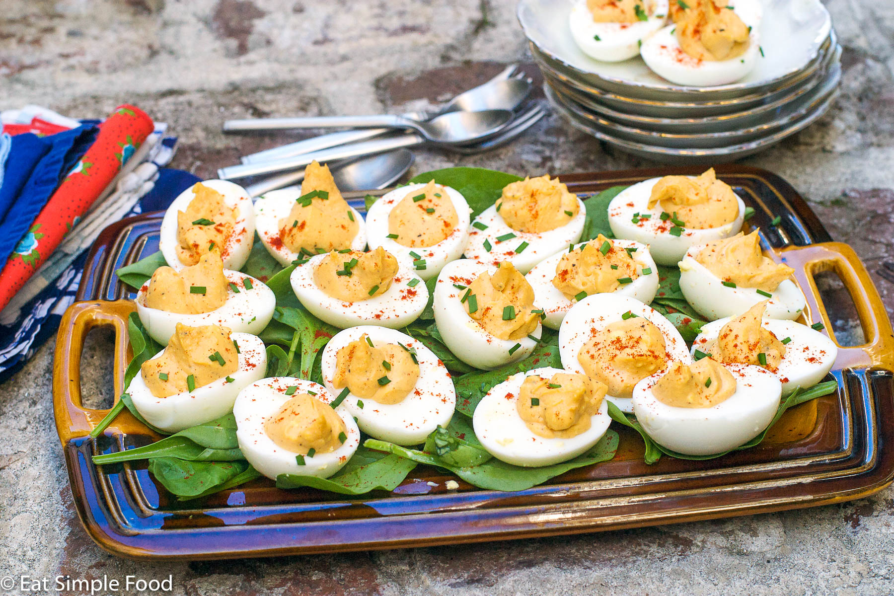 Multiple deviled eggs laying on a bed of spinach on a brown rectangle plate with a chive and paprika garnish.
