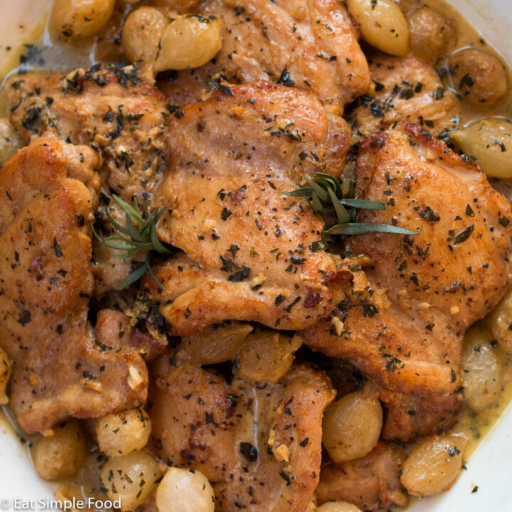 Seared chicken thighs with pearl onions and fresh tarragon with a light brown sauce in a large white bowl with a white napkin with an artichoke on it.