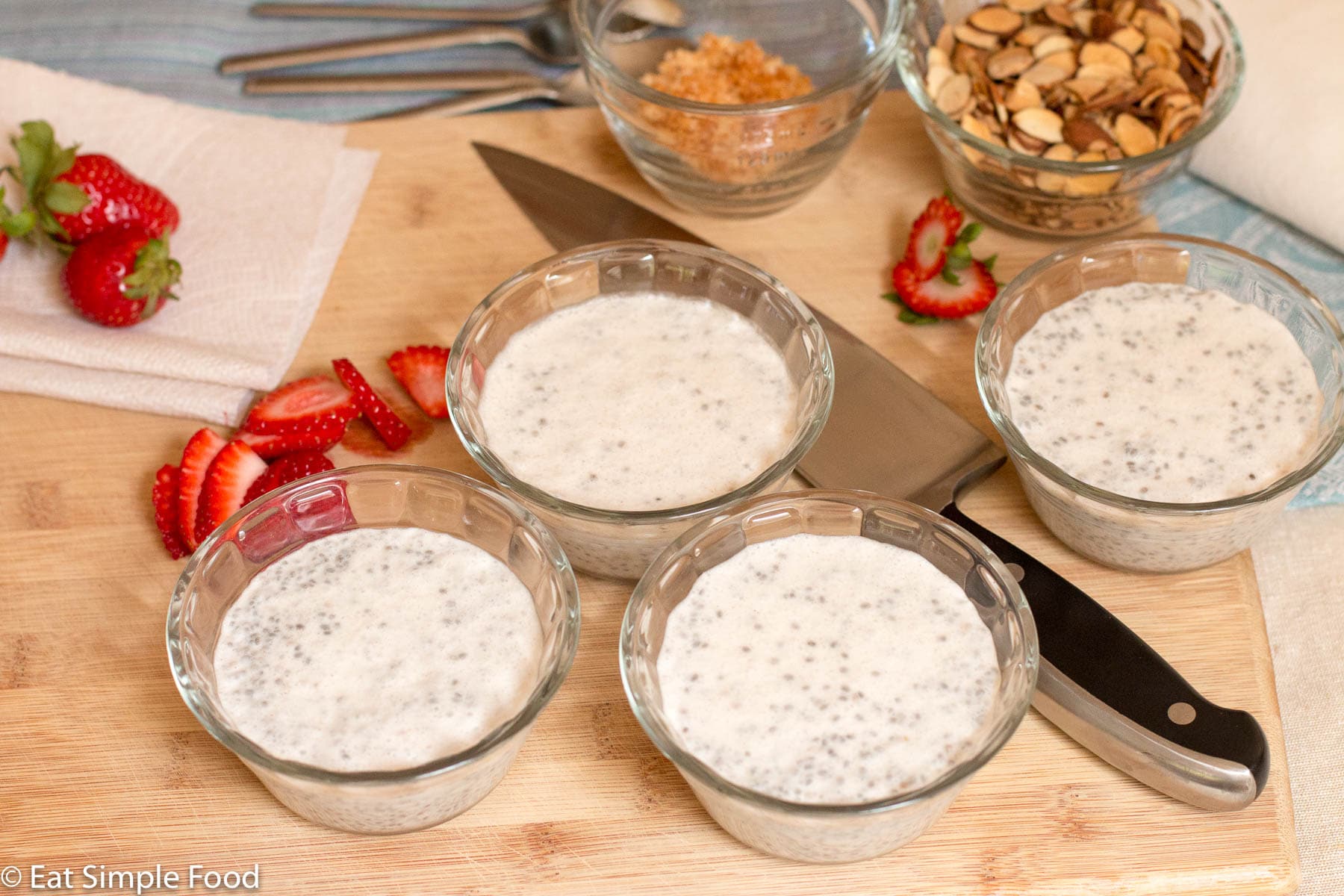 Side view of 4 ramekins filled with white pudding with an assortment of sliced strawberries, toasted coconut flakes, toasted almonds and spoons on the side. On a wood cutting board.