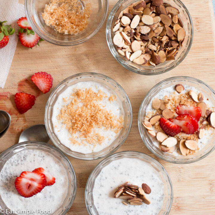 Top down view of 4 ramekins filled with white pudding with an assortment of sliced strawberries, toasted coconut flakes, toasted almonds and spoons. On a wood cutting board.