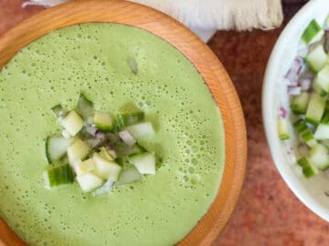Close up of wood bowl of cucumber soup with chunky cucumber and red onion garnish. White bowl of cucumber garnish on side.
