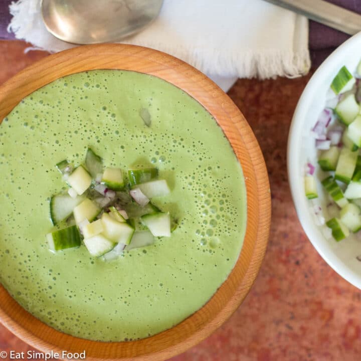 Close up of wood bowl of cucumber soup with chunky cucumber and red onion garnish. White bowl of cucumber garnish on side.