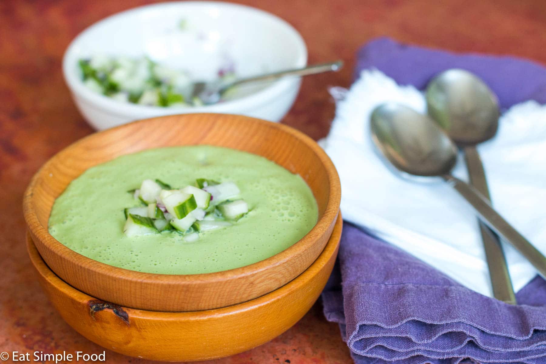 Wood bowl of cucumber soup with chunky cucumber and red onion garnish. White bowl of cucumber garnish on side with small spoon sticking out. 2 spoons on side with purple and white napkins.