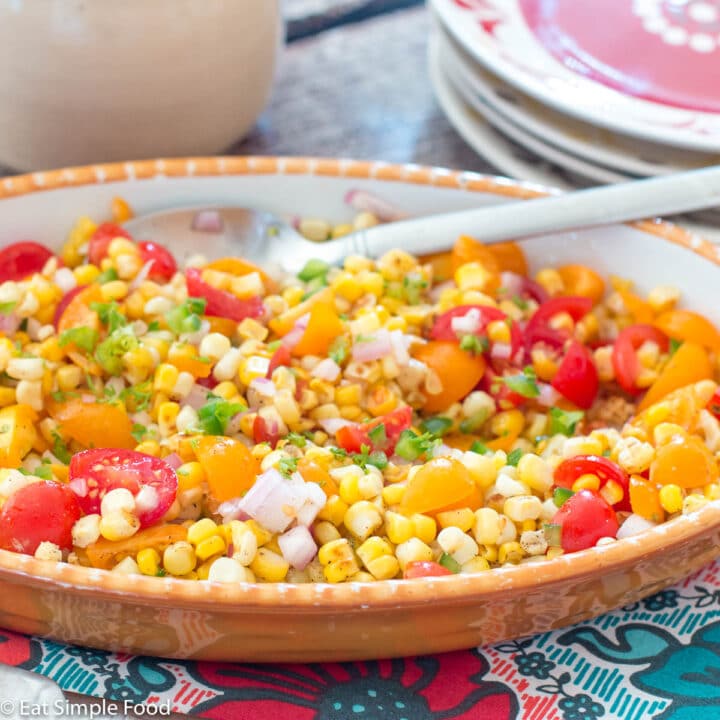 Oval white bowl of corn salad w/ quartered cherry tomatoes, jalapeños, red onions, and cilantro. Side view.