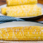 1 white plate with corn on the cob with butter and dusted with chile powder. 3 pieces of plain corn on the cob in the background.