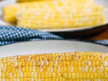 1 white plate with corn on the cob with butter and dusted with chile powder. 3 pieces of plain corn on the cob in the background.