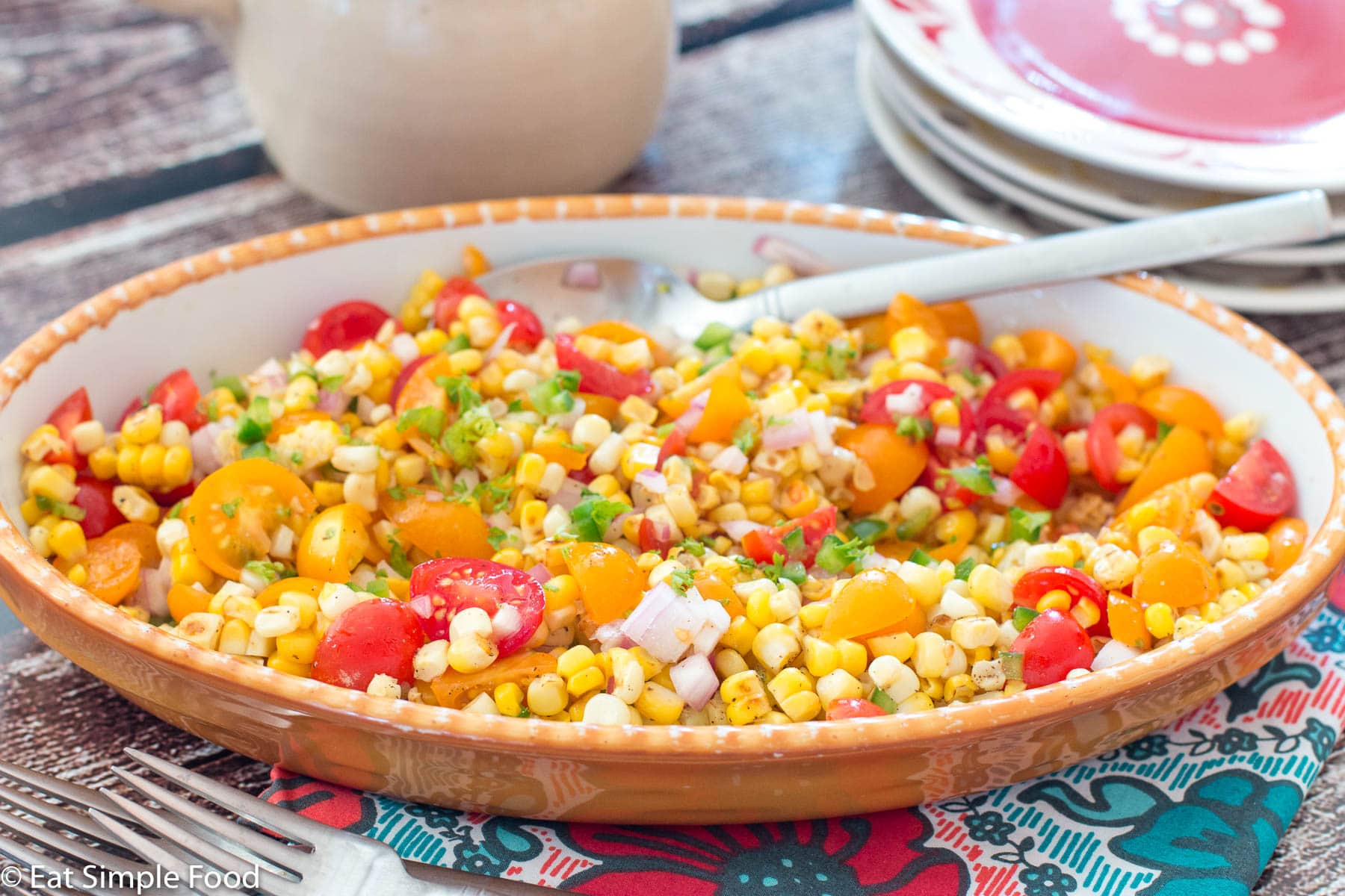 Oval white bowl of corn salad w/ quartered cherry tomatoes, jalapenos, red onions, and cilantro. Side view.