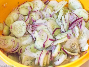 Cucumbers and red onions with dill marinated in vinegar in a large yellow bowl.