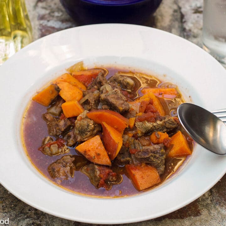 White Bowl With Spoon Filled With Braised Beef and Orange Sweet Potatoes in a Brown Beef Sauce. Side view.