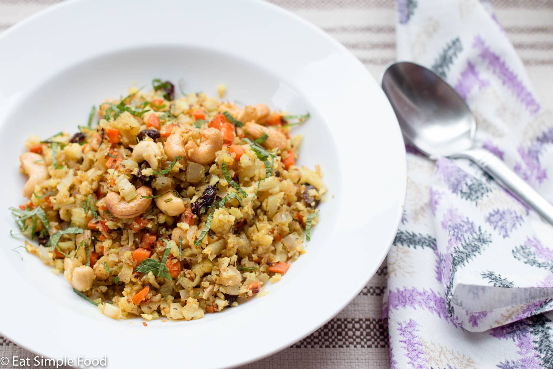 Cauliflower rice with whole cashews, raisins, carrots with curry and garnished with mint. White bowl with spoon. Side view.