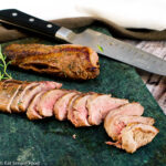 Sliced deer tenderloin on a dark green cutting board with a sprig of rosemary.- Eat Simple Food