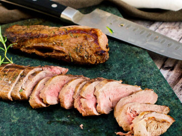 Sliced deer tenderloin on a dark green cutting board with a sprig of rosemary.- Eat Simple Food
