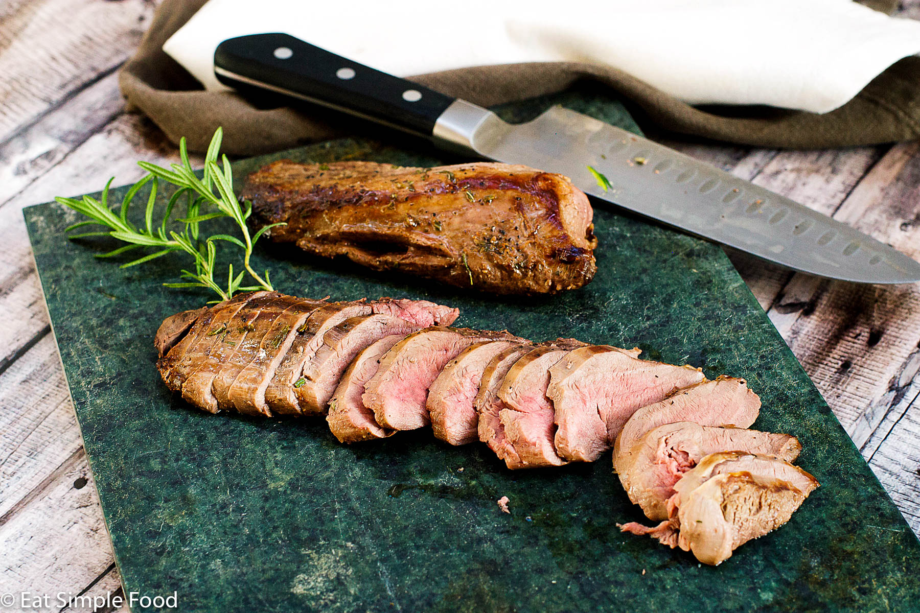 Sliced deer tenderloin on a dark green cutting board with a sprig of rosemary - Eat Simple Food