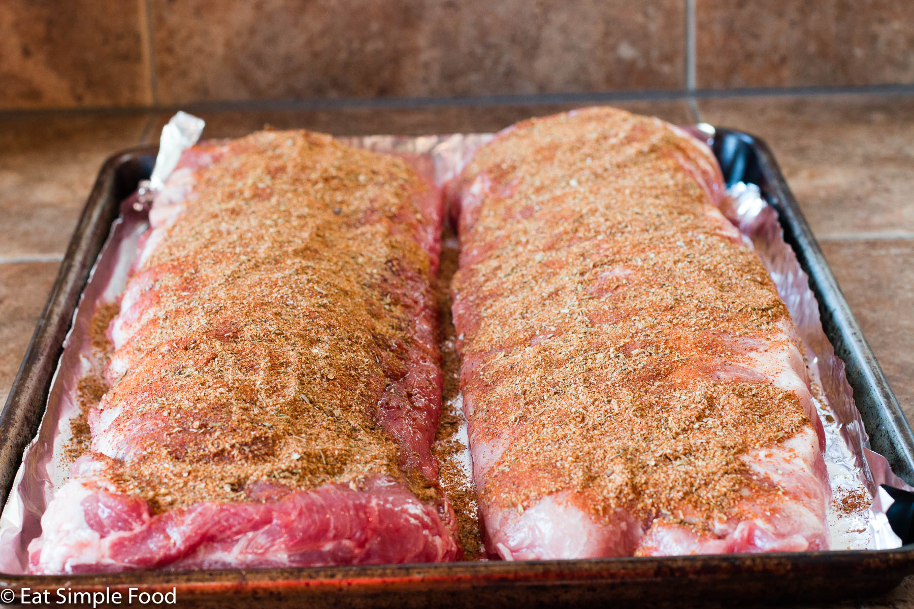 2 racks of baby back ribs on a sheet pan lined with aluminum foil with spices rubbed on the top.