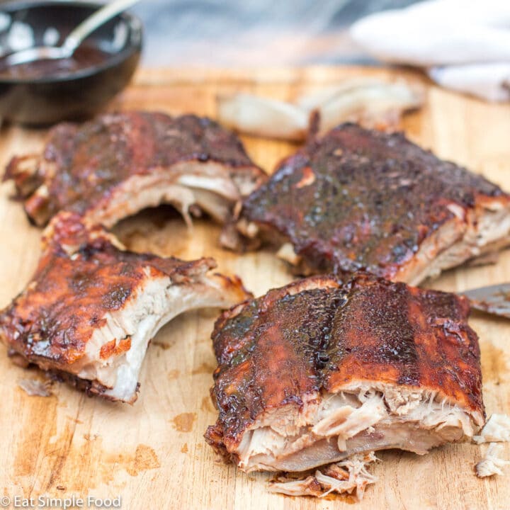 4 pieces (3 ribs each) of Cooked Baby Back Ribs With BBQ Sauce Broiled On with a knife and small black bowl of bbq sauce on a wood cutting board (3 eaten bones in the back). Side view. Horizontal picture.