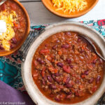 Beef and bean chili in a large brown bowl with a small wood bowl of chili with grated chatter and a spoonful of sour cream.