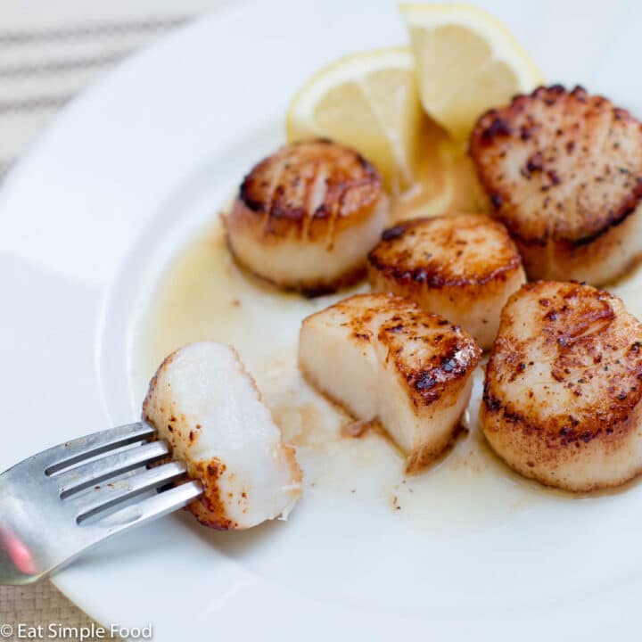 5 scallops on a white plate with 2 lemon wedges. One scallop is cut in half and has a fork sticking in it.