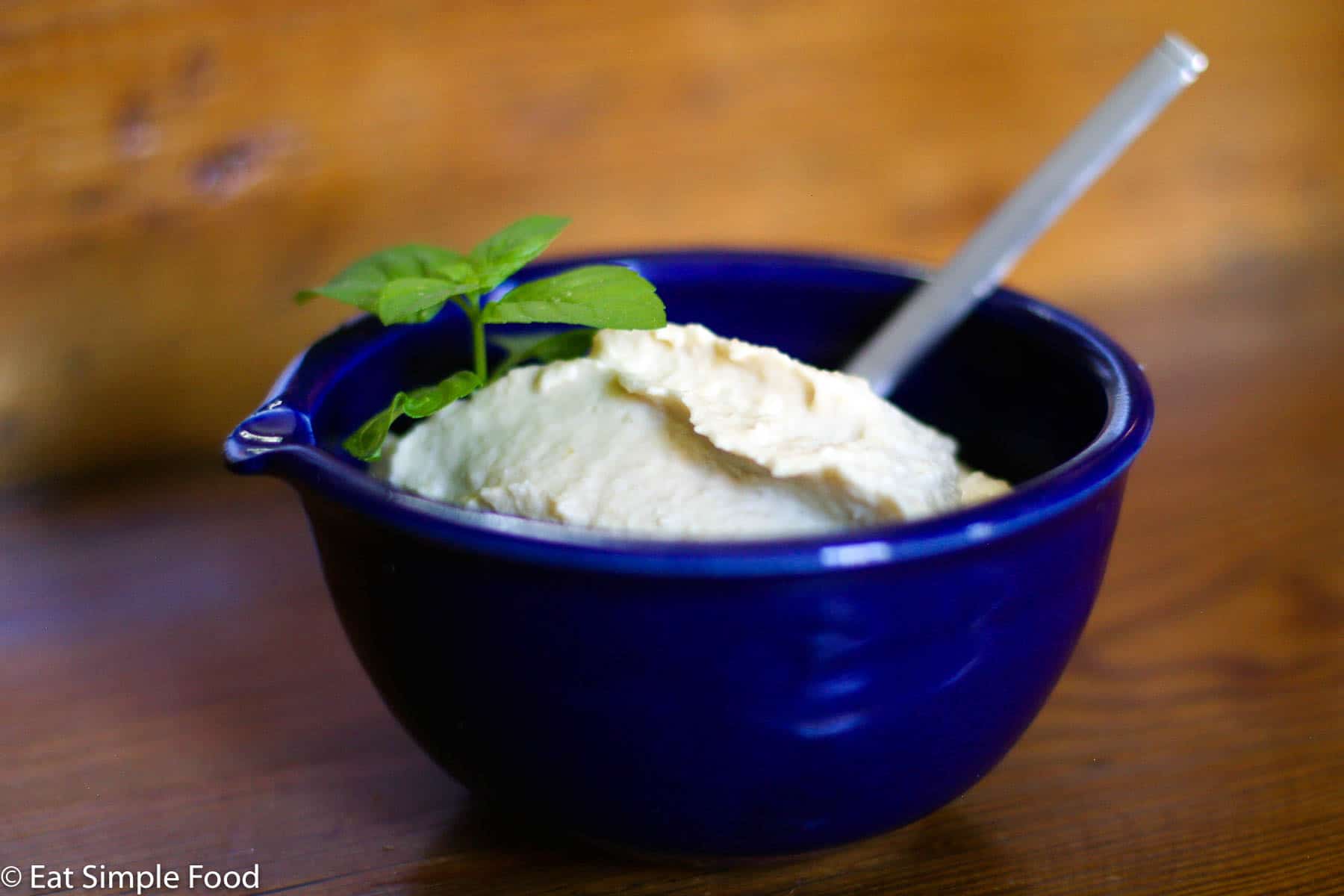 Small blue bowl of off white hummus with a mint garnish. Spoon inserted into hummus.