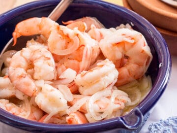 Cooked Pickled Shrimp with thin sliced onions In a blue bowl with a spoon sticking out. 2 wood bowls with small forks on the side. close up.