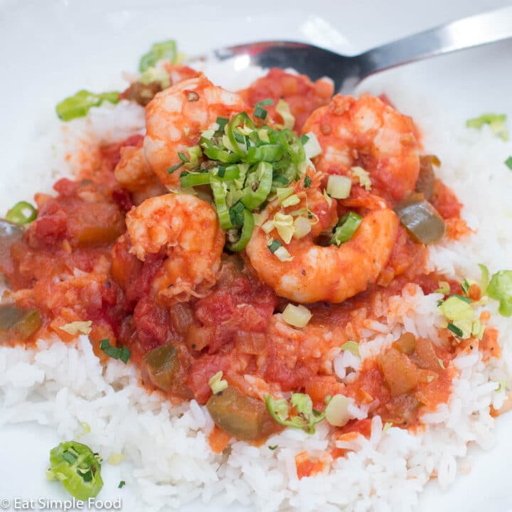 White bowl of shrimp in red tomato sauce over white rice and garnished with peppers and chopped celery leaves.