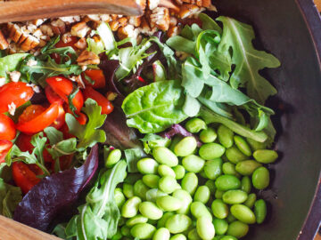 Edamame, Halved cherry Tomatoes, chopped Pecan & Arugula Salad in a wood bowl with wood tongs.