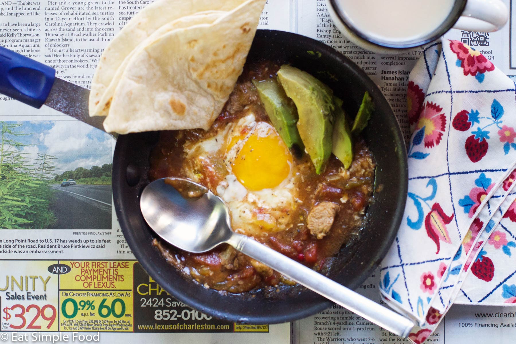 Eggs Baked in Pork Green Chile in a pan with a napkin and a spoon in the pan with a tortilla on the side. Pan laying on an open newspaper. Cup of coffee.