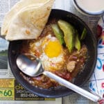 Eggs poached in Pork Green Chile in a pan with a napkin and a spoon in the pan with a tortilla on the side. Pan laying on an open newspaper. Cup of coffee.