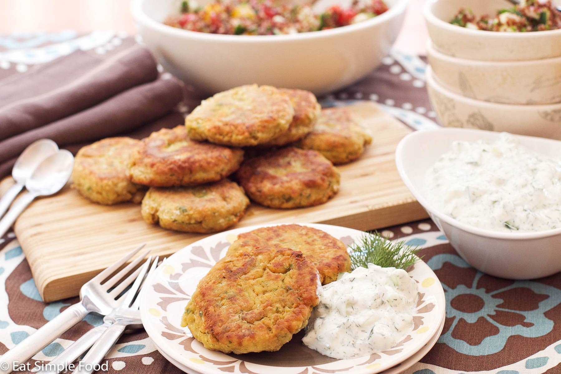 Falafels on a wood cutting board with a side plate of 2 falafels and a bowl of white cucumber tzatziki. side view.