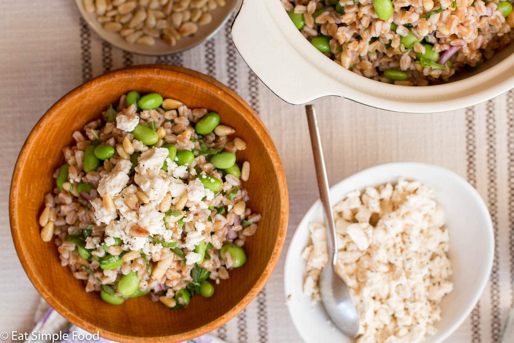 White Bowl of farro, and edamame with a small bowl of toasted pine nuts and another small wood ball with a portion size of the dish topped with feta. top view.