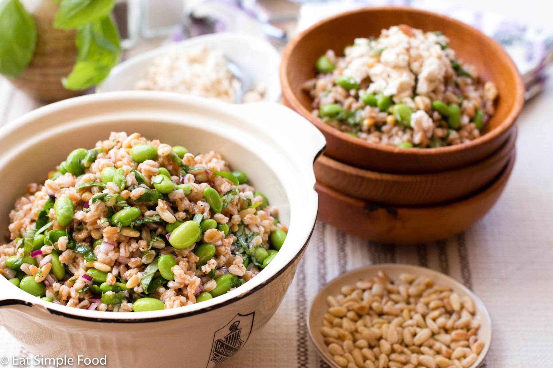 White Bowl of farro, and edamame with a small bowl of toasted pine nuts and another small wood ball with a portion size of the dish topped with feta. side view.
