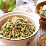 White Bowl of farro, and edamame with a small bowl of toasted pine nuts and another small wood ball with a portion size of the dish topped with feta. side view.